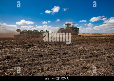 Provinz Nordkasachstan, Kasachstan - 12. Mai 2012: Frühjahrsaat. John Deere Traktor kultiviert Boden mit Pflug in Staubwolke. Blauer Himmel, s Stockfoto