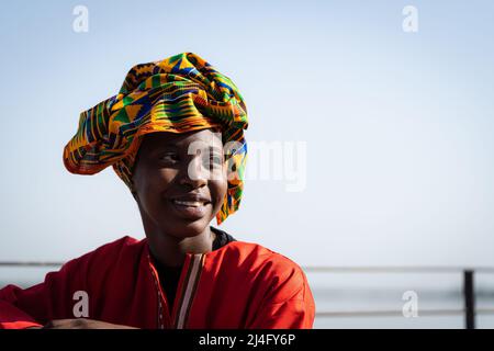 Porträt einer strahlenden afrikanischen Schönheit, die einen roten Kaftan und ein farbenfrohes Kopftuch mit traditionellem Muster trägt, das lässig am Fluss sitzt Stockfoto