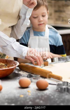 Mutter und Sohn schneiden auf einem schwarzen Holztisch, der zu Hause in der Küche mit Mehl bestreut ist, Formen zum Backen von süßen Plätzchen aus Blätterteig aus. Auswahl Stockfoto