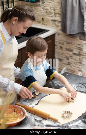 Mutter und Sohn schneiden auf einem schwarzen Holztisch, der zu Hause in der Küche mit Mehl bestreut ist, Formen zum Backen von süßen Plätzchen aus Blätterteig aus. Auswahl Stockfoto