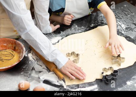 Mutter und Sohn schneiden auf einem schwarzen Holztisch, der zu Hause in der Küche mit Mehl bestreut ist, Formen zum Backen von süßen Plätzchen aus Blätterteig aus. Auswahl Stockfoto