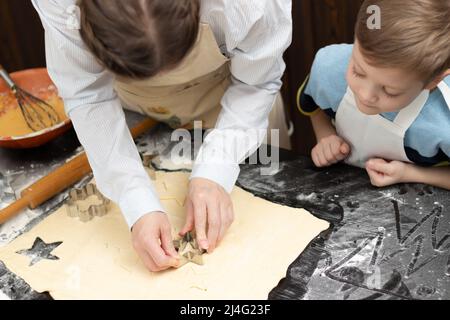 Mutter und Sohn schneiden auf einem schwarzen Holztisch, der zu Hause in der Küche mit Mehl bestreut ist, Formen zum Backen von süßen Plätzchen aus Blätterteig aus. Auswahl Stockfoto