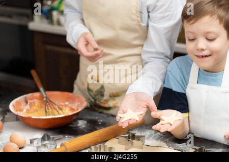 Mutter und Sohn schneiden auf einem schwarzen Holztisch, der zu Hause in der Küche mit Mehl bestreut ist, Formen zum Backen von süßen Plätzchen aus Blätterteig aus. Auswahl Stockfoto