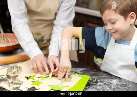 Mutter und Sohn schneiden auf einem schwarzen Holztisch, der zu Hause in der Küche mit Mehl bestreut ist, Formen zum Backen von süßen Plätzchen aus Blätterteig aus. Auswahl Stockfoto