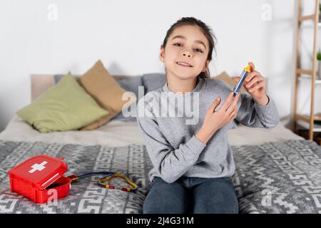 Lächelndes Mädchen, das eine Spielzeugspritze hält, während es auf dem Bett neben dem erste-Hilfe-Set und dem Stethoskop sitzt Stockfoto