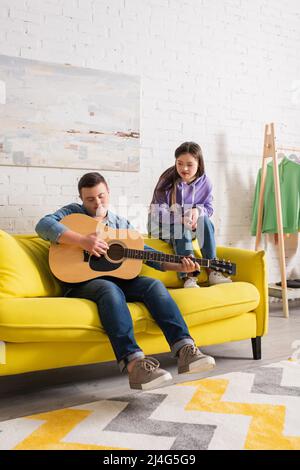 Teenager mit Down-Syndrom spielt akustische Gitarre in der Nähe von Freund zu Hause Stockfoto
