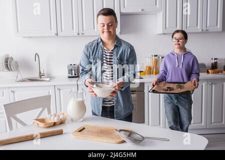 Teenager mit Down-Syndrom hält Mehl in der Nähe Freundin mit Backblech in der Küche Stockfoto