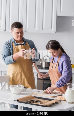 Teenager mit Down-Syndrom Kochen Teig in der Nähe Freund mit Smartphone zu Hause Stockfoto