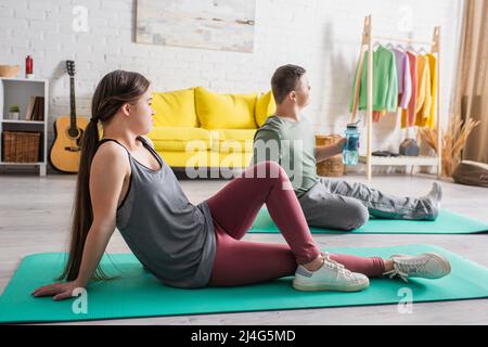 Seitenansicht eines Teenagers mit Down-Syndrom, der auf einer Fitnessmatte in der Nähe eines Freundes zu Hause sitzt Stockfoto
