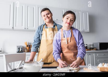 Fröhliches Paar mit Down-Syndrom, das beim Kochen in der Küche die Kamera anschaut Stockfoto