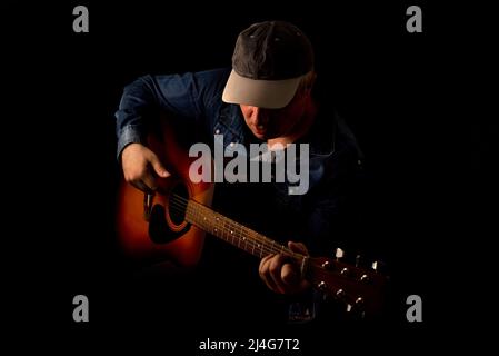 Session-Gitarrist in einem Jeans-Hemd spielt eine akustische Gitarre auf schwarzem Hintergrund. Low-Key-Hochformat. Stockfoto