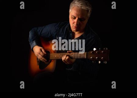 Session-Gitarrist in einem Jeans-Hemd spielt eine akustische Gitarre auf schwarzem Hintergrund. Low-Key-Hochformat. Stockfoto