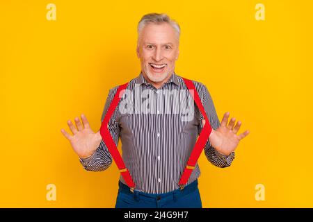 Portrait von attraktiven fröhlich erstaunt lustig grauhaarigen Mann ziehen Hosenträger mit Spaß isoliert über glänzen gelben Hintergrund Stockfoto