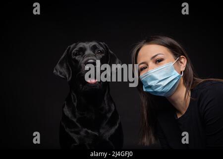 Porträt eines Labrador Retriever Hundes in einer schützenden medizinischen Maske mit einer weiblichen Besitzerin. Das Bild wurde in einem Fotostudio aufgenommen Stockfoto