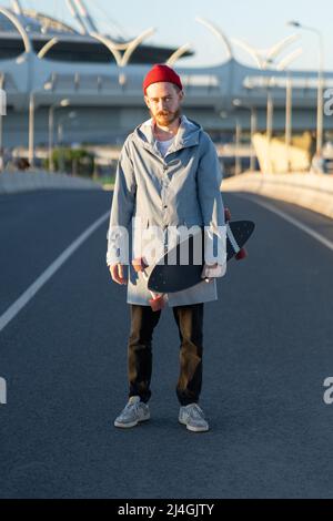 Junger bärtiger Hipster in rotem Hut hält Longboard in der City Street. Trendiges urbanes Lifestyle-Konzept Stockfoto