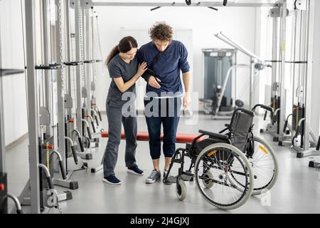 Reha-Spezialist hilft einem Kerl aus einem Rollstuhl stehen Stockfoto