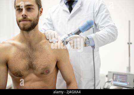 Der Arzt wendet eine Schockwellentherapie auf die Schulter des Mannes an Stockfoto