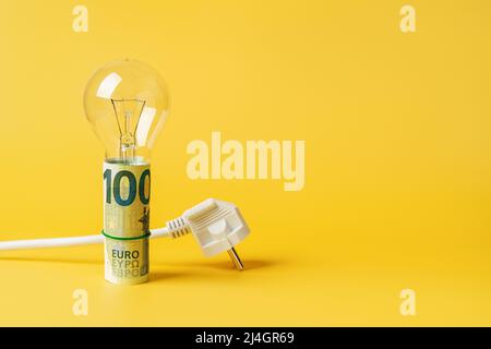 Glühbirne auf gerollten 100-Euro-Banknoten und weißer Netzstecker auf gelbem Hintergrund. Speicherplatz kopieren. Konzept der Erhöhung der Stromkosten. Stockfoto