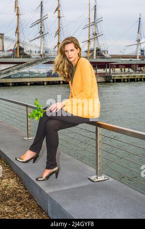 Eine junge, hübsche Frau, die grüne Pflanzen hält, sitzt auf einem Zaun am Fluss und denkt tief nach. Hintergrund ist ein Hafen. Stockfoto