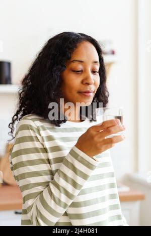 Lächelnde afroamerikanische Frau, die auf einem weichen fokussierten Hintergrund ein Glas klares Wasser anschaut Stockfoto
