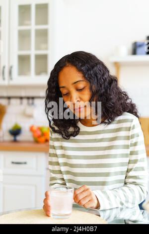 Erschöpfte afroamerikanische Frau wartet darauf, dass sich die Pille in einem Glas Wasser in der Küche auflöst Stockfoto