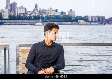 18 Jahre alter ostindischer amerikanischer Teenager, der sich draußen in New York City entspannt, schwarzes Langarmshirt trägt, auf einem Stuhl am East River sitzt, lo Stockfoto