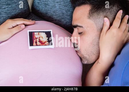 Nahaufnahme Mann, der auf dem Bett liegt und den Bauch seiner Schwangeren küsst, mit dem gedruckten Ultraschallfoto auf dem Bauch. Stockfoto