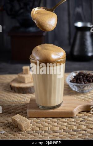 Iced Dalgona Coffee, trendiger, flauschiger, cremiger Kaffee auf dunklem Holzhintergrund mit Kaffeemühle, Zucker und Kaffeebohnen, mit Schaumlöffel Stockfoto