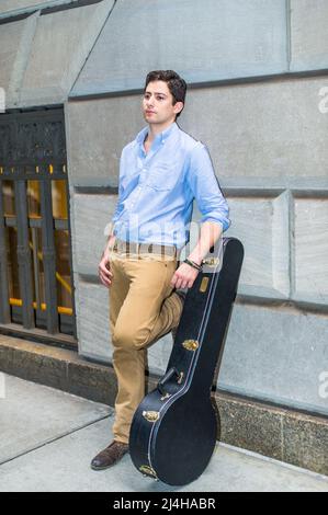 Ein junger Musiker mit einer Musikinstrumentenbox steht auf der Straße und lehnt sich an die Wand, in Gedanken verloren. Stockfoto