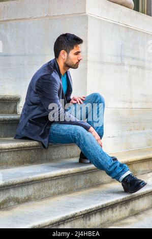 Ein junger, gutaussehender Kerl mit einem kleinen Brot und Schnurrbart sitzt auf einer Treppe vor einem Bürobui in einem dunkelvioletten Wollblazer und einer blauen Jeans Stockfoto