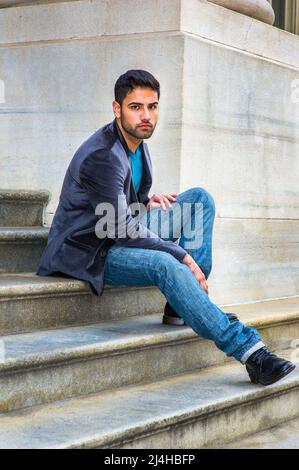 In einem dunkelvioletten Wollblazer, einer blauen Jeans und schwarzen Lederschuhen sitzt ein junger, hübscher Kerl mit ein wenig Brot und Schnurrbart auf der Stufe Stockfoto