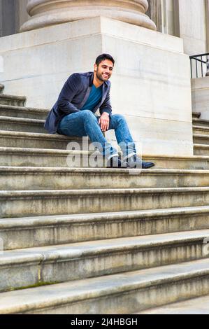 Ein junger Mann aus dem Nahen Osten mit Brot und Schnurrbart sitzt auf einer Treppe, bekleidet mit einem dunkelvioletten Wollblazer, blauen Jeans und schwarzen Lederschuhen, Stockfoto