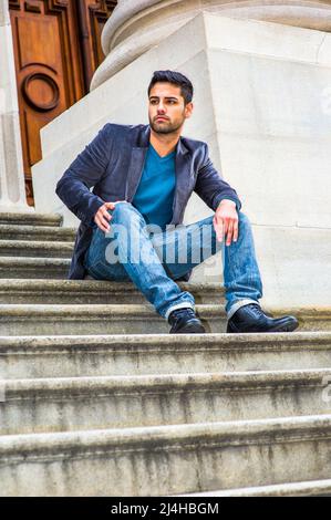 Ein junger Mann aus dem Nahen Osten mit Brot und Schnurrbart sitzt in einem dunkelvioletten Wollblazer, einer blauen Jeans und schwarzen Lederschuhen auf der Treppe i Stockfoto