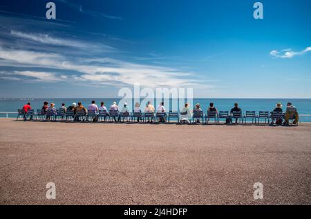 Die Menschen sitzen auf den berühmten blauen Stühlen am Ufer in Nizza - Frankreich, sie beobachten azurblaues Wasser des Ligurischen Meeres, Ruhe Stockfoto