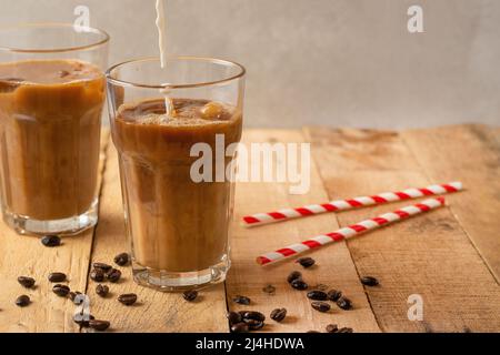 Kalter Kaffee, der in die Creme eingießt, Milch in den durchsichtigen Gläsern mit Eis und den Trinkhalmen, auf dem hölzernen Hintergrund, das kühlende Getränk, erfrischend Stockfoto