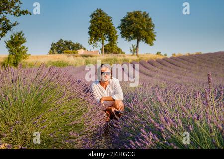 Der gutaussehende, brutale Mann mit langen, brünetten Haaren sitzt auf dem Lavendelfeld in der provence in der Nähe von Valensole, Frankreich, bei klarem sonnigem Wetter, in einer Reihe von Stockfoto