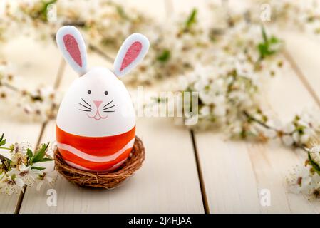 Osterei mit niedlichem Hasengesicht und Hasenohren neben blühenden Frühlingsblumen auf einem Holztisch. Osterkonzept. Stockfoto