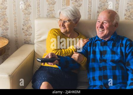 Lachend gealtertes kaukasisches Paar, Mann und Frau eine Comedy-Show beim Fernsehen, während sie auf einem beigefarbenen Ledersofa sitzen. Gemütliche, altmodische Einrichtung. Hochwertige Fotos Stockfoto