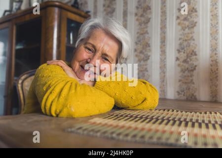 Die alte, grauhaarige Rentnerin in einem gelben Pullover, die ihre Arme auf einem Holztisch ruhte, die Kamera ansah und friedlich lächelte. Altmodische Innenausstattung. Hochwertige Fotos Stockfoto