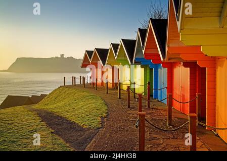 Großbritannien, North Yorkshire, Scarborough, farbenfrohe Strandhütten über der North Bay Promenade mit Sonnenaufgang über Scarborough Castle Stockfoto