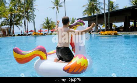 Lustiger Mann, der Spaß hat, auf schwimmenden aufblasbaren Einhorn tanzt, tropischer Urlaub Stockfoto