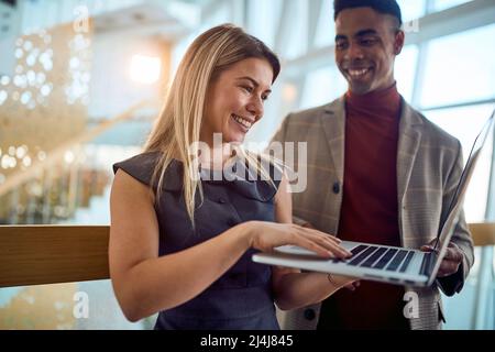 Zwei junge, fröhliche Geschäftsleute nutzen einen Laptop, während sie in einer angenehmen Atmosphäre auf dem Flur stehen. Unternehmen, Mitarbeiter, Unternehmen Stockfoto