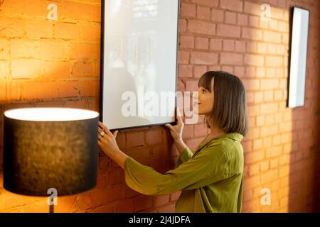 Eine Frau hängt zu Hause einen Fotorahmen an einer Ziegelwand Stockfoto