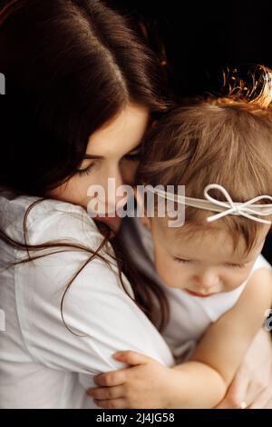 Dunkelhaarige junge Mutter hält sanft in ihren Armen und umarmt kleine Tochter auf schwarzem Hintergrund, glücklich fürsorgliche Mutter umarmen lull kleines Baby Mädchen Kind Stockfoto