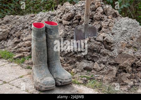 Eine Schaufel in einem Haufen Tonerde, nachdem man ein Stück Erde von Hand gegraben und schmutzige grüne Stiefel bekommen hat Stockfoto