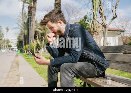 Der junge Mann trägt eine blaue Jeansjacke und erhält am Telefon schlechte Nachrichten, Misserfolge und Krisenkonzept. Sitzen auf einer Parkbank im Stadtpark. Allein, traurig und Stockfoto
