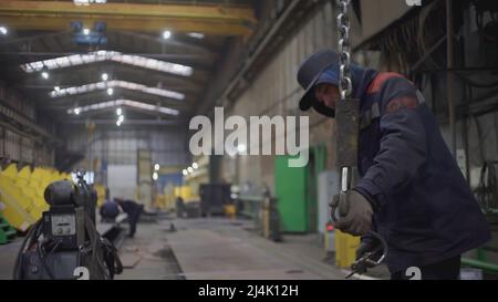 MOSKAU, RUSSLAND - 25. FEBRUAR 2022 : Ein Mann in einer Fabrik hakt Ausrüstung. CLIP. Maschinenantrieb auf einem Metallhaken in einem Industriegebäude. Ein Mann in der Arbeit Stockfoto