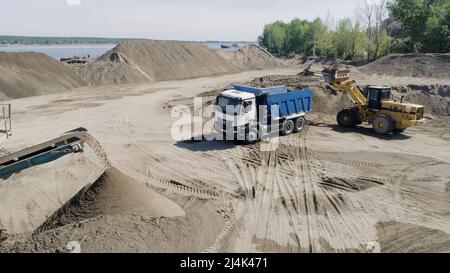 Der Bauprozess. Szene. Lastwagen transportieren Baumaterialien in einem leeren Gebiet, hinter dem sich ein Fluss, Wälder und blauer Himmel befinden Stockfoto