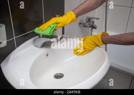 Hände in gelben Handschuhen reinigen Waschbecken und Wasserhahn mit Mikrofasertuch. Housekeeping- und Hygienekonzept Stockfoto