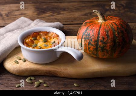 Kürbiskäse-Auflauf in Keramikform mit Kürbiskernen und frischem Kürbis über Holzstruktur-Hintergrund, Freiraum, horizontal Stockfoto